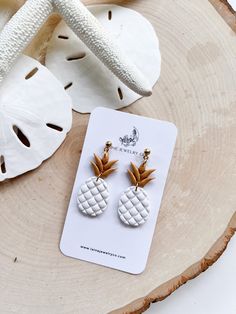 the pineapple earrings are on display next to a white piece of wood and some seashells