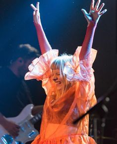 a woman in an orange dress on stage with her arms up and hands raised above her head