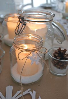 three mason jars filled with white candles and snow flakes on a table next to pine cones