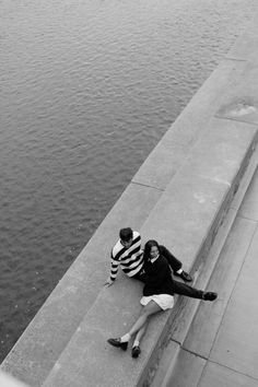 black and white photograph of two people sitting on the edge of a pier next to water