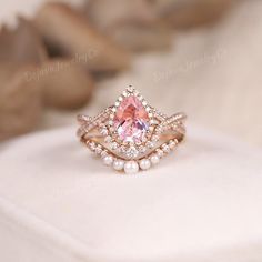 a pink diamond and pearl ring sitting on top of a white cushioned surface with other jewelry items in the background