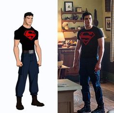 the man is wearing a superman t - shirt and jeans, while standing in front of a table