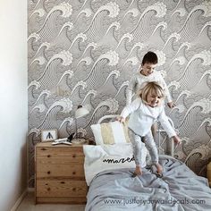 two children jumping on a bed in front of a wallpapered room with waves