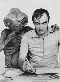 a man sitting at a table with an alien doll next to him and writing on a piece of paper