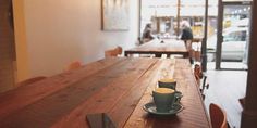 two cups of coffee sit on a wooden table in front of a window with people walking by