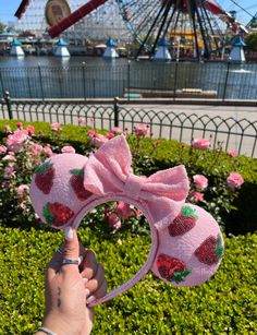 a hand holding up a pink minnie mouse ears with strawberries on it and a ferris wheel in the background