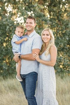 a man and woman holding a baby in their arms while standing in tall grass with trees behind them