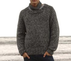 a man standing on the beach with his hands in his pockets and wearing a sweater