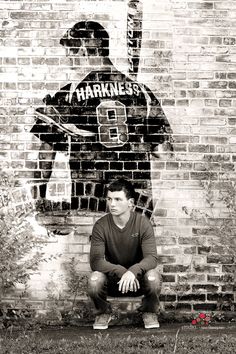 a black and white photo of a man sitting in front of a brick wall with a painting of a baseball player on it