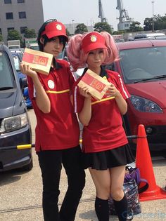 two women in red uniforms holding pizza boxes and posing for the camera with cars behind them