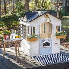 a small white doll house sitting on top of a wooden deck next to trees and bushes