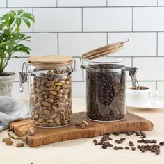 two glass jars filled with coffee beans sitting on top of a wooden cutting board next to a potted plant