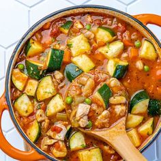 an orange pot filled with vegetable stew on top of a white tile counter next to a wooden spoon