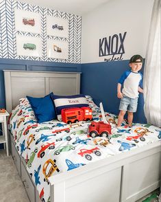 a young boy standing on top of a bed next to a toy train and truck