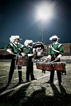 three men in green and white uniforms are playing drums on the football field at night