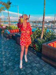 a woman in a red dress is holding a drink and looking at the water with palm trees behind her