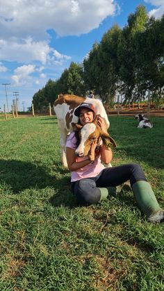 a woman sitting on the grass holding a dog and a cow in front of her