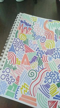 a spiral notebook with colorful doodles on the cover sitting on a table next to a coffee cup