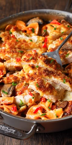 a skillet filled with chicken and vegetables on top of a wooden table next to a fork