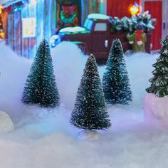 three small christmas trees in the snow near a red truck and building with lights on it