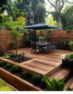 a wooden deck with chairs and tables surrounded by trees, plants and an umbrella in the middle