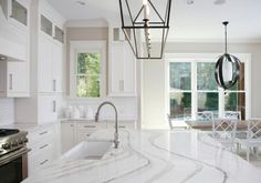 a white kitchen with marble counter tops and an island in front of a window that looks out onto the backyard