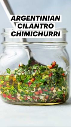 a glass jar filled with greens and other vegetables on top of a white table next to a spoon