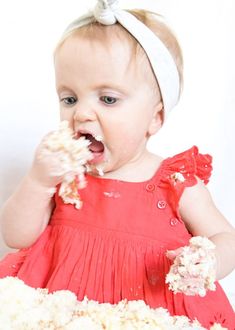 a baby girl in a red dress is eating some food from her hands and wearing a white headband