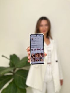 a woman holding up a cell phone in front of her face while standing next to a potted plant