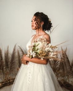 a woman in a wedding dress holding a bouquet and looking off into the distance with tall grass behind her