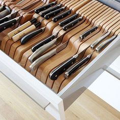 a drawer with many knives in it on top of a wooden floor next to a white table