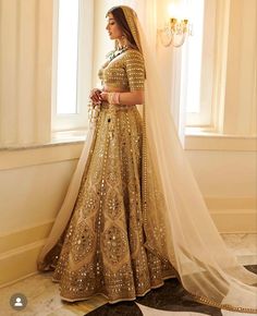 a woman in a gold and white bridal gown standing by a window with her veil pulled back