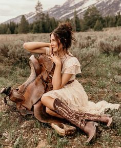 a woman sitting on the ground next to a horse in a field with mountains in the background