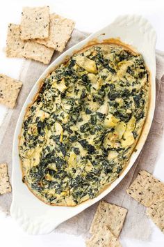 spinach and cheese dip in a white dish surrounded by crackers on a table