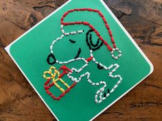 a snoopy christmas ornament on a green square with red, white and yellow beads