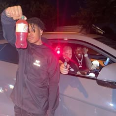 a man holding up a red bottle in front of a car with another person behind him