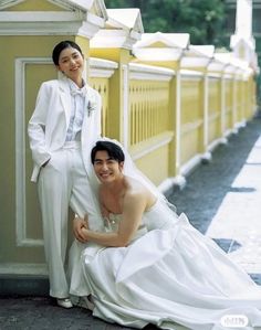 a man and woman posing for a photo in front of a yellow wall wearing white clothes