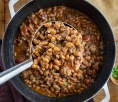 a ladle full of beans and meat in a pot on top of a wooden table