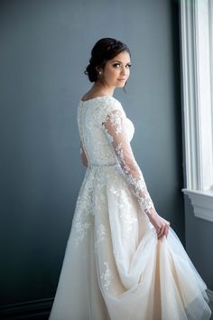 a woman in a wedding dress standing by a window with her hands on her hips