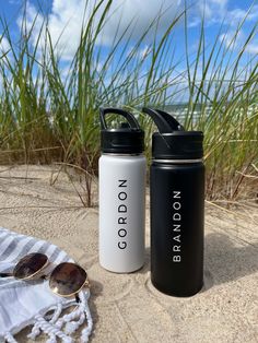 two black and white water bottles sitting on the sand next to sunglasses, towel and beach grass