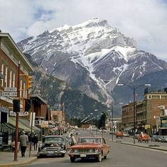 an old car is driving down the street in front of a mountain with snow on it