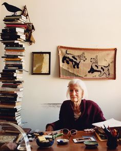 an old woman sitting at a table with many books in front of her and a cat on the wall behind her