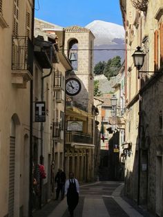 two people walking down an alley way with buildings on both sides and a clock tower in the distance