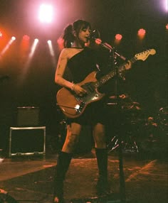 a woman in black dress playing guitar on stage with lights and microphones behind her