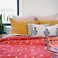 a bed with orange and pink sheets, pillows and plants in the window sill