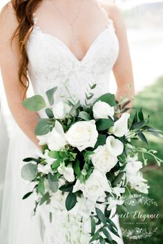 the bride is holding her bouquet with white flowers and greenery in her hand while standing outside