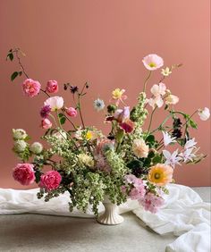 a vase filled with lots of flowers on top of a white cloth covered table next to a pink wall