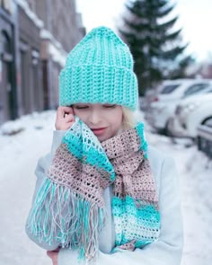 a woman wearing a hat and scarf standing in the snow with her hands on her head