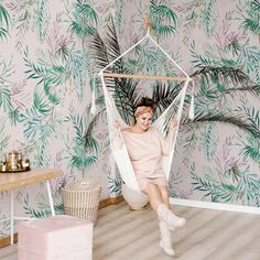 a woman sitting in a hammock with palm leaves on the wall behind her