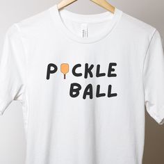 Introducing our new pickleball shirt, perfect for both on and off the court! Made from a lightweight fabric, this shirt will keep you comfortable no matter how long your pickleball game lasts. The relaxed fit ensures that you have full range of motion, allowing you to move freely and play your best. This shirt features a classic crew neck design and short sleeves, making it versatile enough to wear on any occasion. The shirt also features a fun and colorful design with a pickleball theme, showing off your love for the sport. The high-quality print will stay vibrant even after many washes, ensuring that you can enjoy wearing this shirt for years to come. This casual pickleball shirt is available in a range of sizes to fit every body type. It's perfect for both men and women and can be dress White Tennis Top With Team Name, White Moisture-wicking Tennis T-shirt, White Moisture-wicking T-shirt For Tennis, Pickleball Graphic Tee With Screen Print, Team Spirit T-shirt For Pickleball With Short Sleeves, Short Sleeve Tops With Team Name For Pickleball, White T-shirt With Team Name For Pickleball, Sporty White T-shirt For Pickleball, Pickleball Shirts